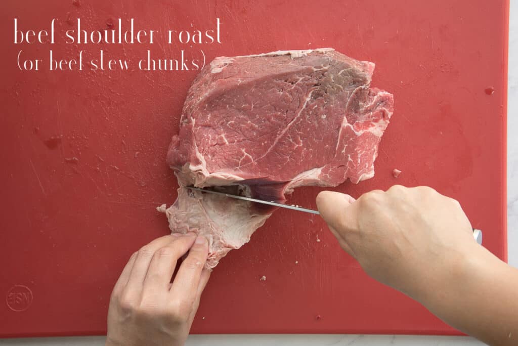 The beef shoulder roast is trimmed of excess fat on a red cutting board. White text overlay says beef shoulder roast (or beef stew chunks).