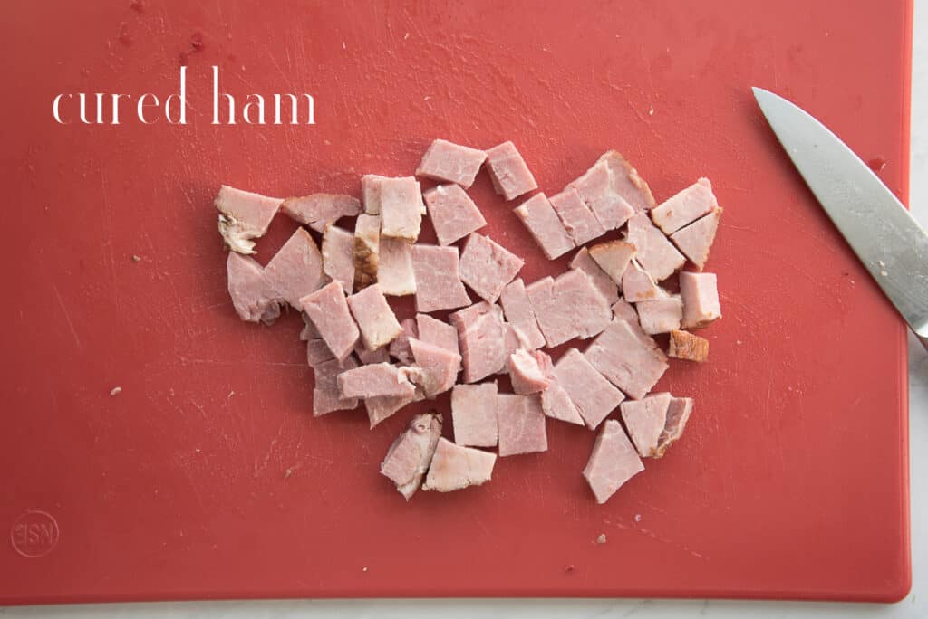Diced cured ham on a red cutting board with the knife used to cut it. White text overlay says cured ham.