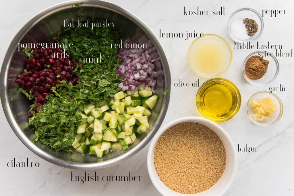The ingredients to make Tabbouleh are shown: pomegranate arils, parsley, red onion, mint, English cucumber, cilantro in a silver mixing bowl. Next to the bowl are: kosher salt, lemon juice, olive oil, bulgur, garlic, Middle Eastern Spice Blend, and pepper.