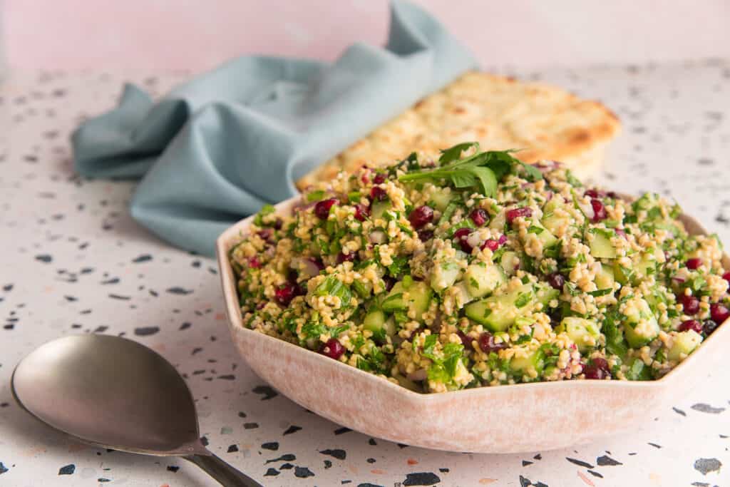 Horizontal sideview of a pink bowl filled with Tabbouleh next to a silver serving spoon.