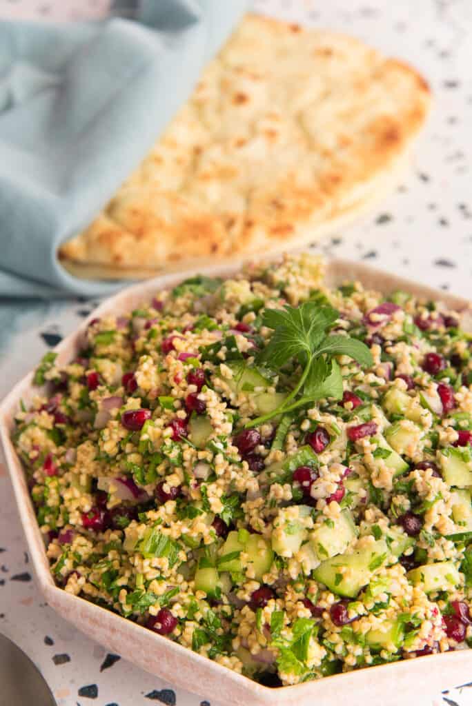 Portrait image of a pink bowl of Tabbouleh garnished with a sprig of parsley. Naan wrapped in a blue kitchen towel is in the top left background.