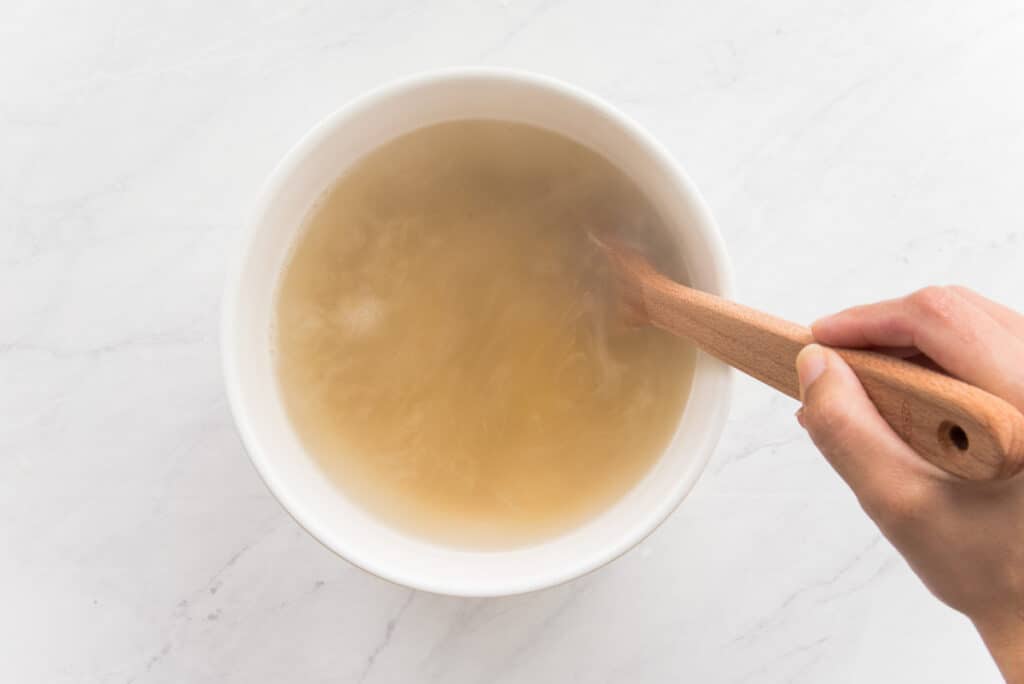 A hand uses a wooden spoon to stir boiling water and bulgur together in a white bowl.