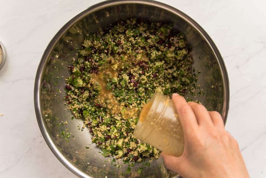 The lemon vinaigrette is poured from a glass jar over the tabbouleh in a silver mixing bowl.