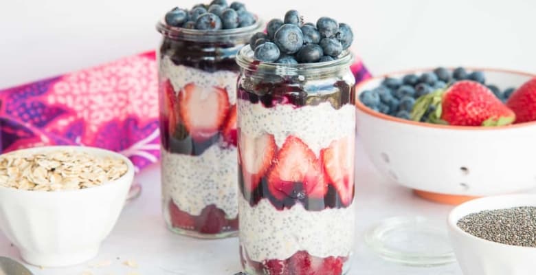 Horizontal image of two tall weck jars filled with Chia Oats Fruit Parfaits. White bowls of oats, chia seeds, and fresh fruit surround the jars.