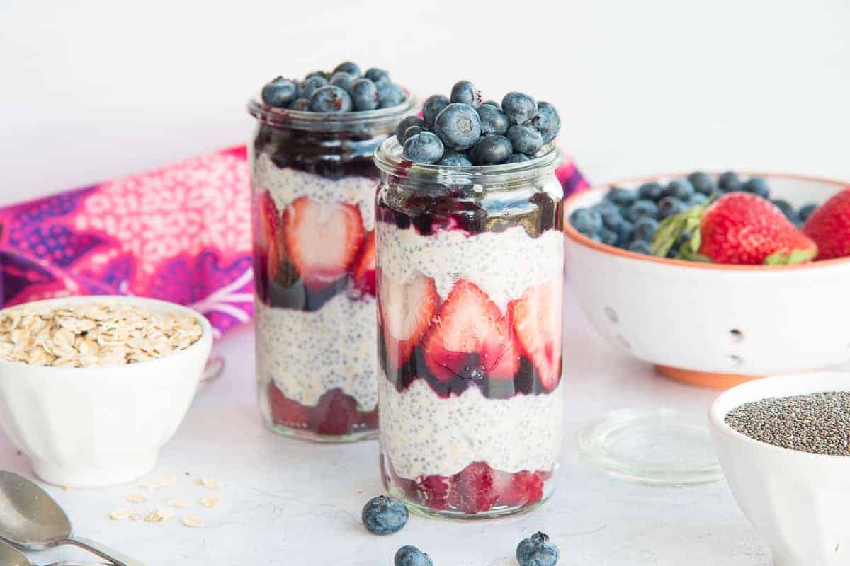 Horizontal image of two tall weck jars filled with Chia Oats Fruit Parfaits. White bowls of oats, chia seeds, and fresh fruit surround the jars.