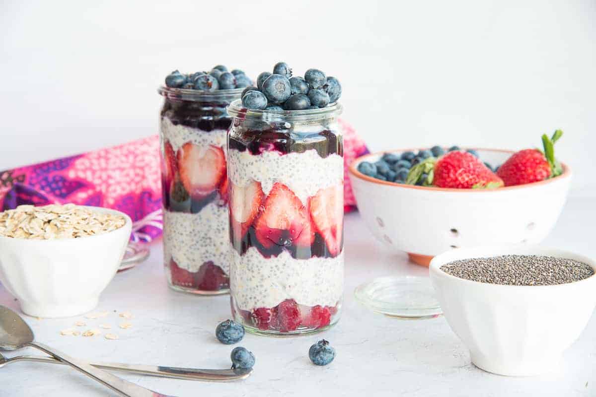 Horizontal image of two clear glass weck jars with Chia Oats Fruit Parfaits layered. White bowls of oats, chia seeds, and berries surround the jars.
