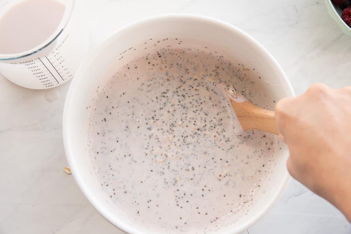 A wooden spoon stirs the chia oats and kefir together in a white ceramic mixing bowl.
