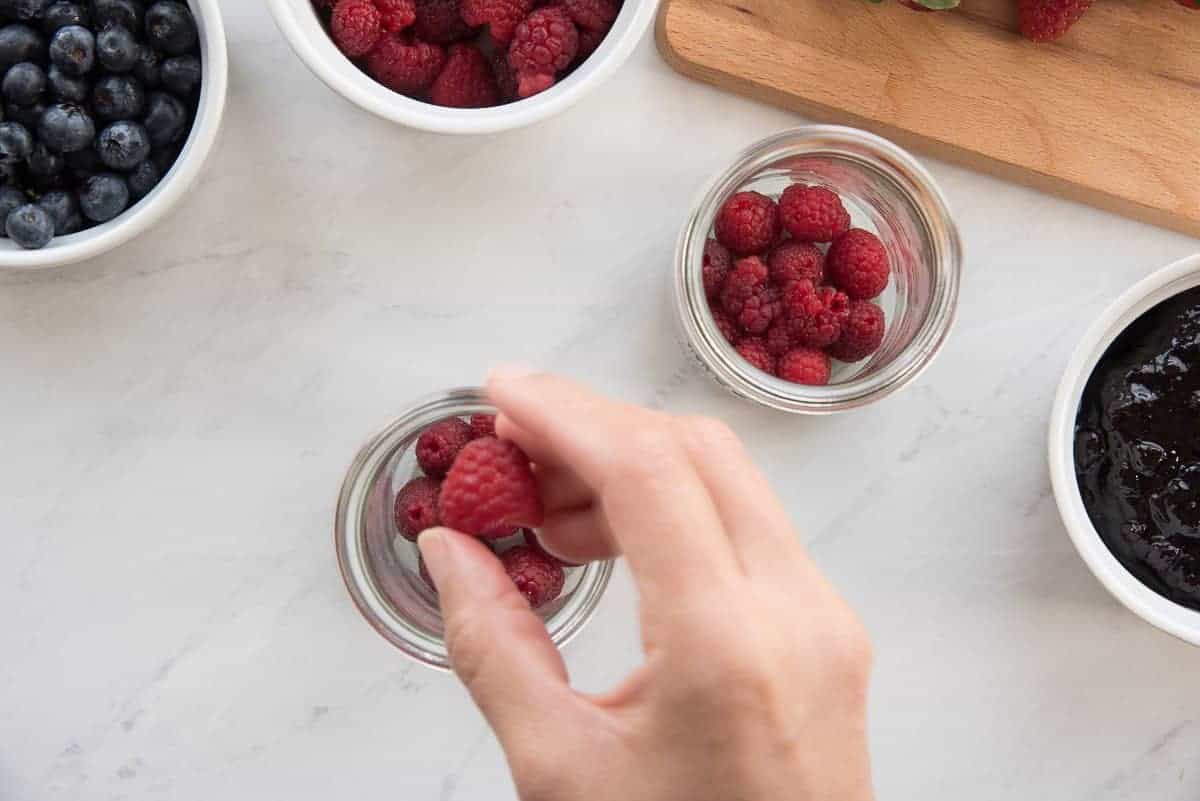 Hand adds raspberries to glass jars to begin the parfaits.