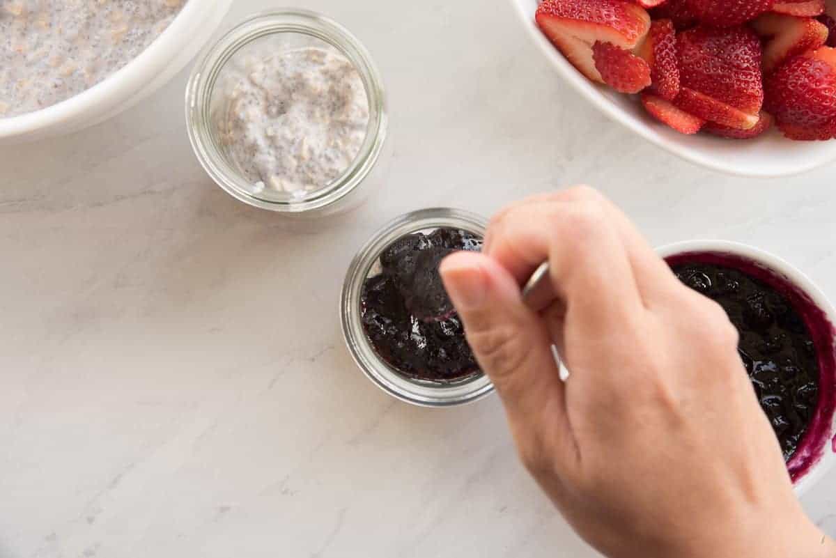 A hand uses a long handled spoon to add fruit preserves to the pudding in a tall glass jar.