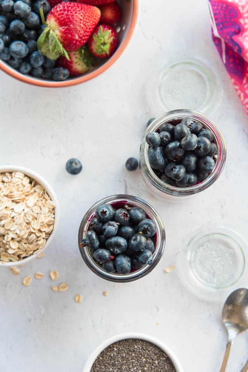 Overhead portrait image of two jars with blueberries on top. Surrounding the jars are fresh berries, and bowls of oats and chia seeds.