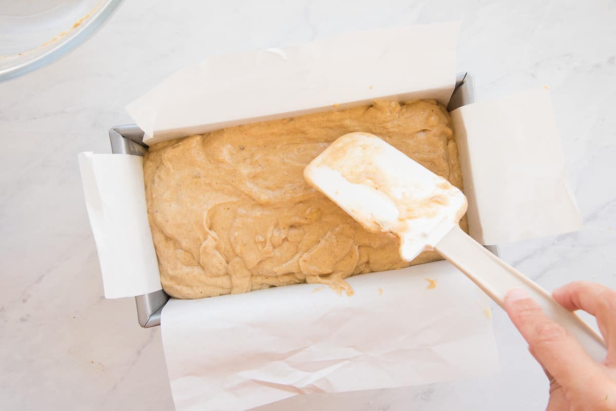 A rubber spatula smooths the batter in a silver loaf pan lined with parchment paper.