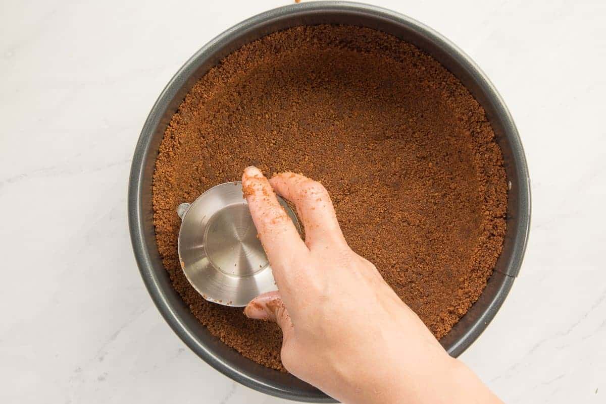 A hand uses a measuring cup to press the graham crust into a pan.