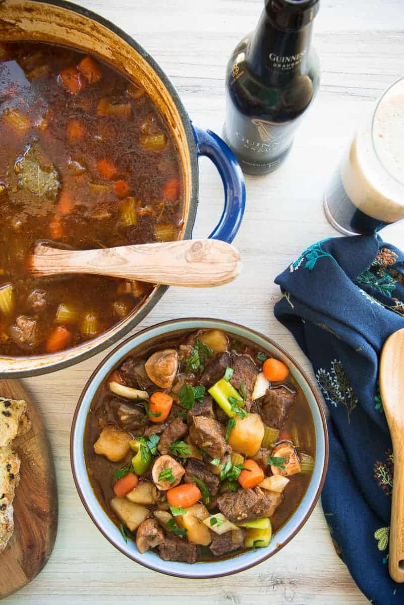 Guinness Beef stew in a blue bowl next to a blue pot of stew and a bottle of guinness poured into a glass.