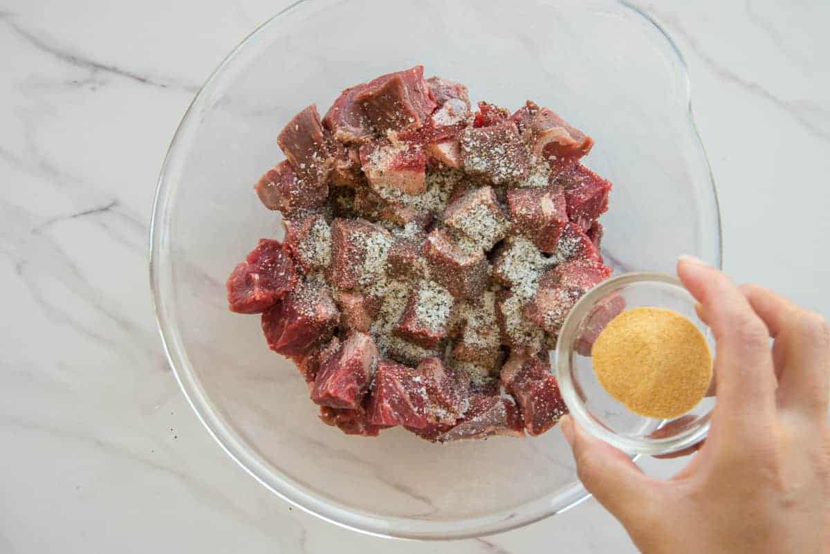 Garlic powder is added to seasoned beef chunks in a glass mixing bowl.