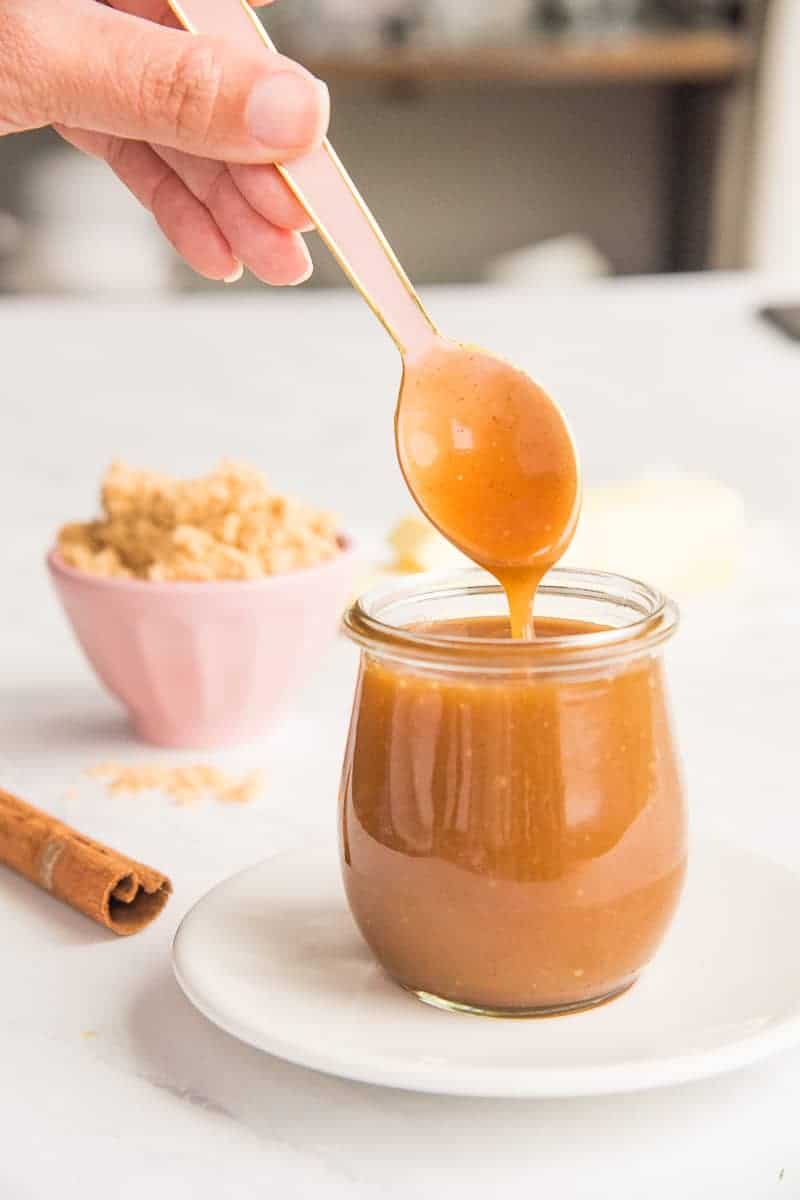 Pink spoon with Spiced Toffee Sauce dripping from it into a glass jar.