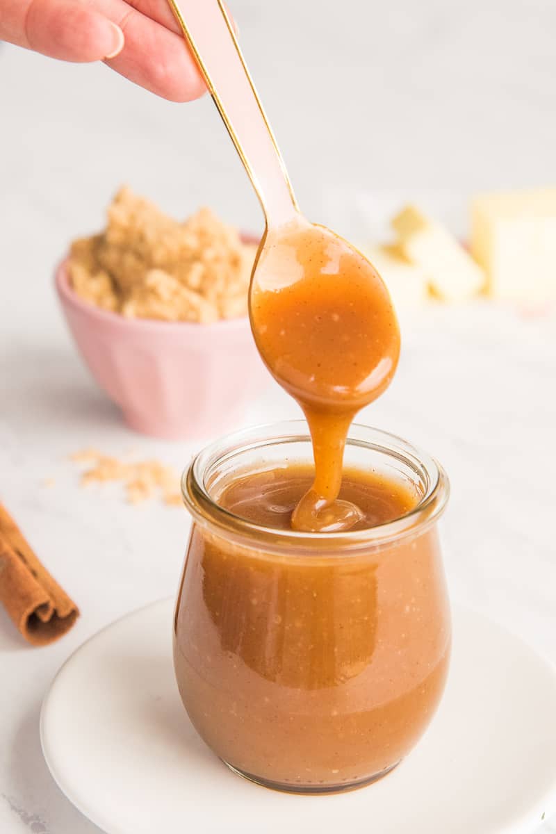 A hand holds a pink spoon that's dripping Spiced Toffee Sauce into a glass jar.