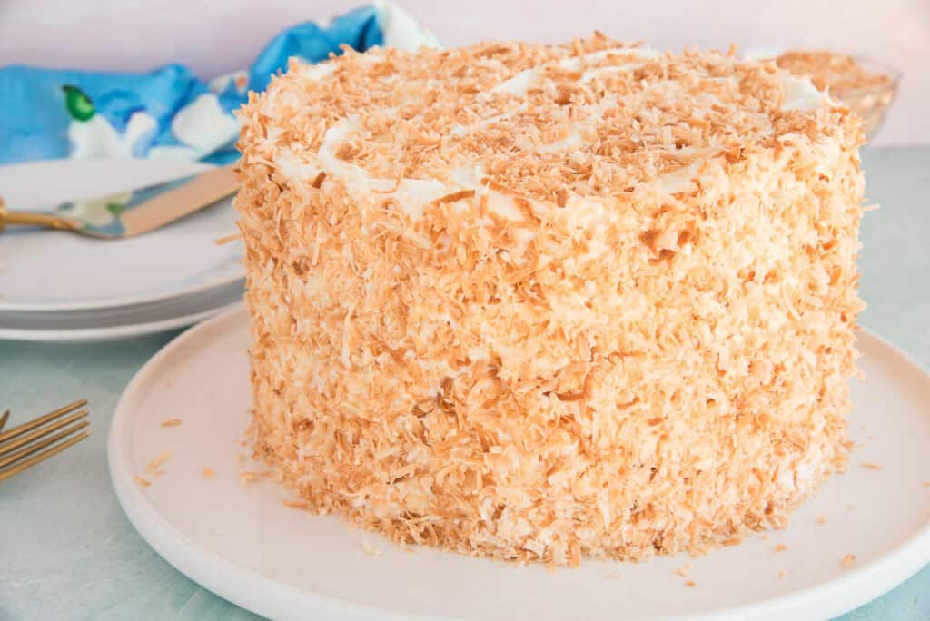A round cake covered in crispy toasted coconut flakes sits on a white plate. In the background, there are stacked white plates, a gold knife and fork, and a blue and white cloth.