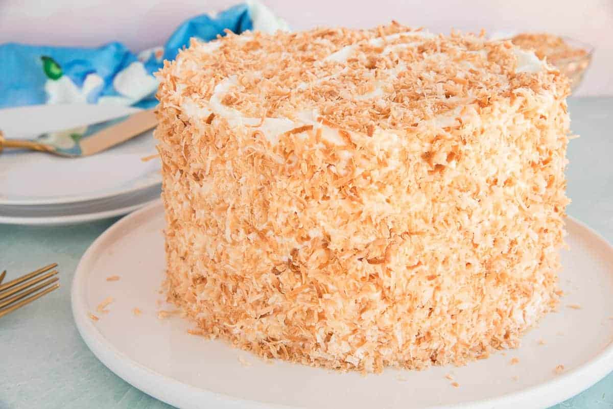 A whole Toasted Coconut Cake on a white cake plate garnished with toasted coconut flakes.