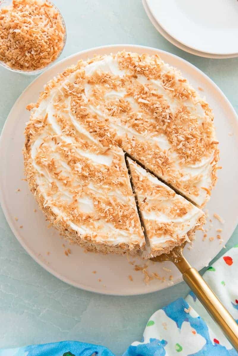 A slice of Toasted Coconut Cake is getting ready to be pulled from the whole cake on a white plate.
