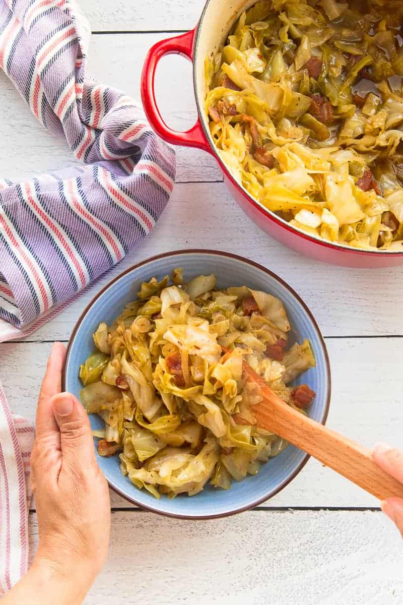 A hand holds on to a blue bowl while spooning the Braised Cabbage with Bacon into it.