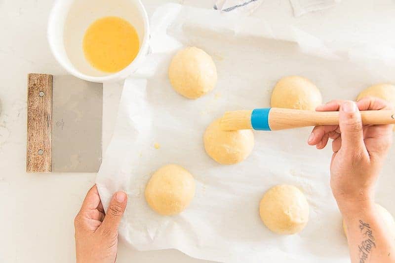 A hand uses a pastry brush to glaze the Brioche Rolls with egg wash.
