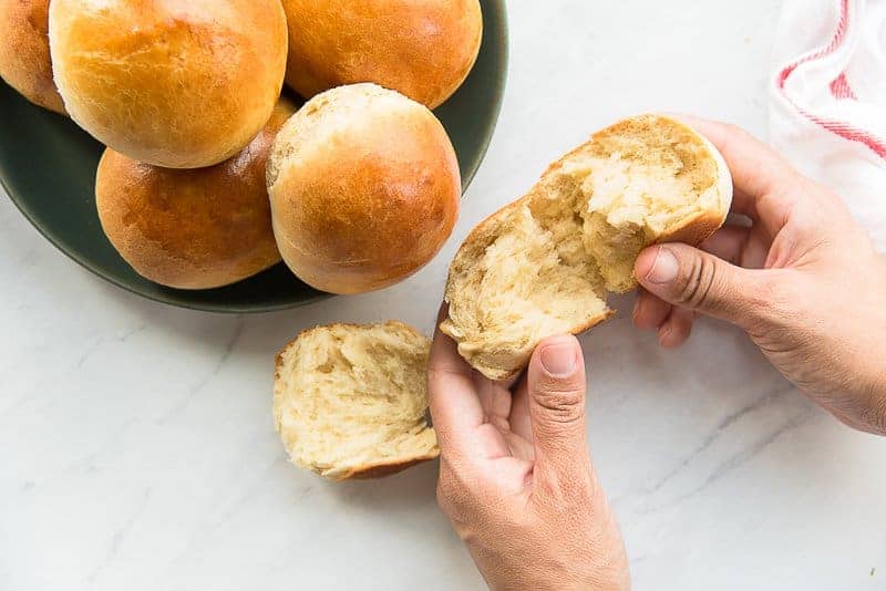 Hands tear open a Brioche Roll to show the fluffy interior. A green bowl of Brioche Rolls is next to it.