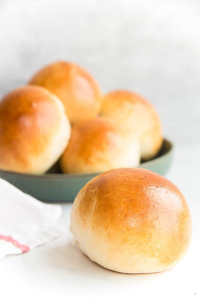 A single Brioche Roll is in the right foreground in front of a dark green bowl of Brioche Rolls.