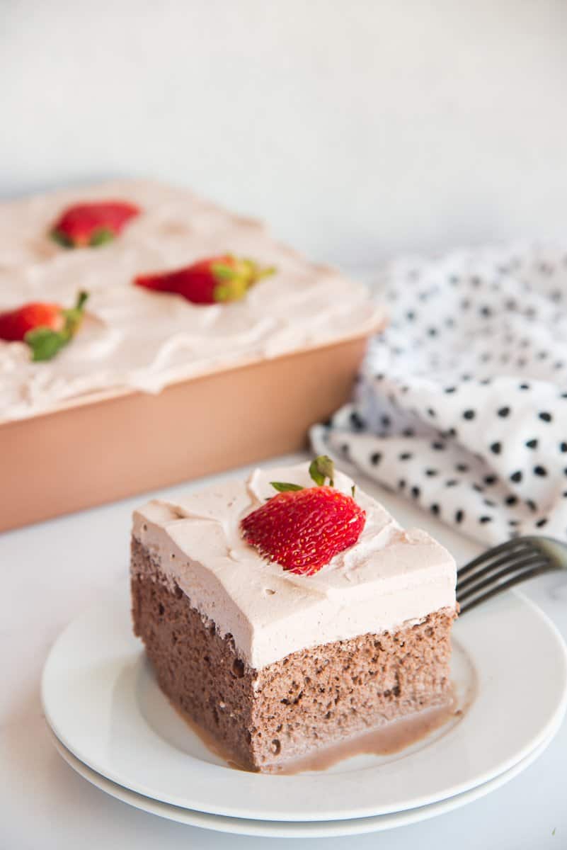 A slice of Chocolate Tres Leches Cake on a white plate is garnished with a fanned strawberry.