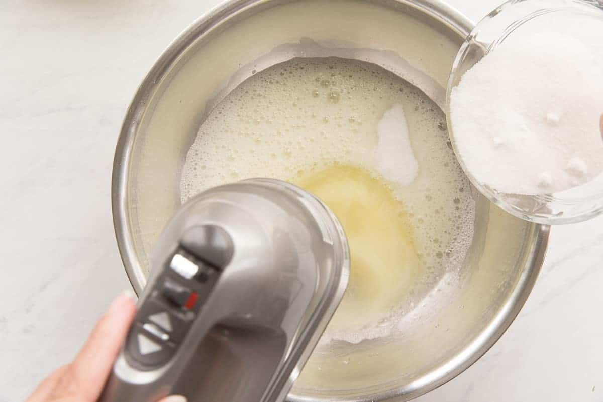 Sugar is gradually added to the egg whites in a metal bowl as they're being whipped by an electric hand mixer.