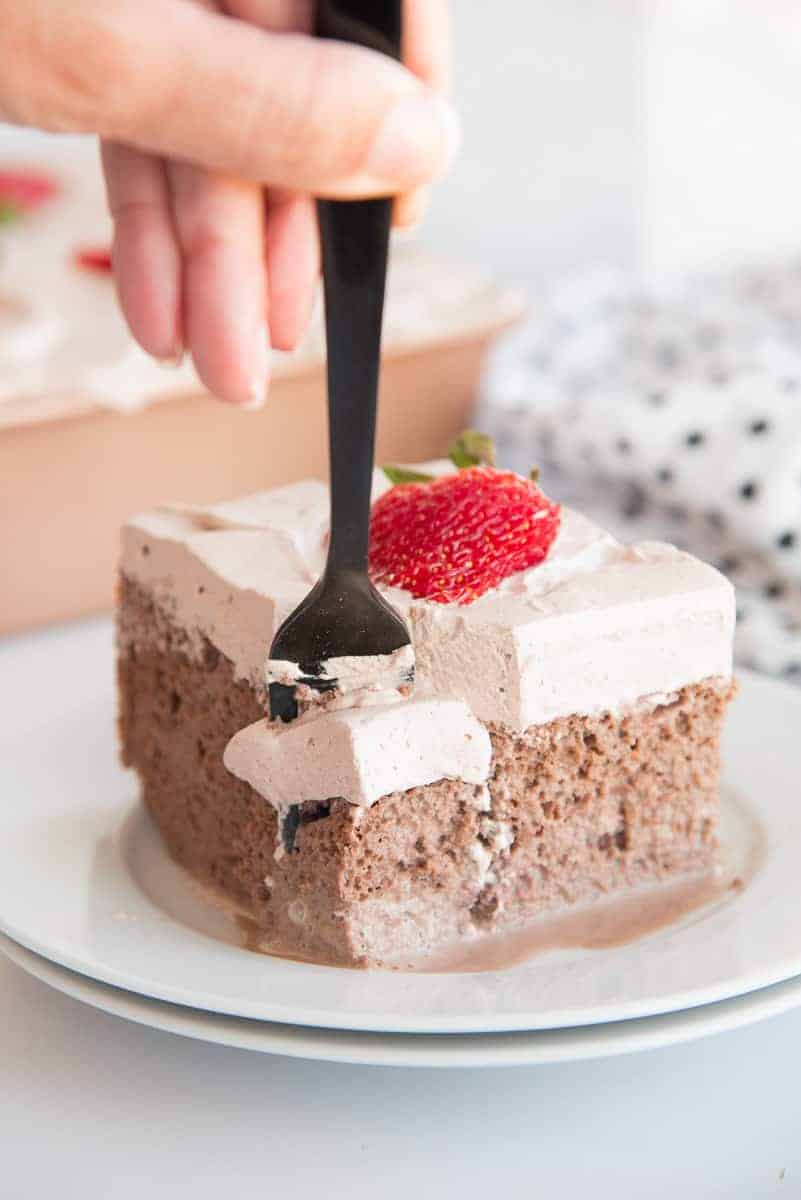 A hand pushes a black fork down into a slice of Chocolate Tres Leches Cake