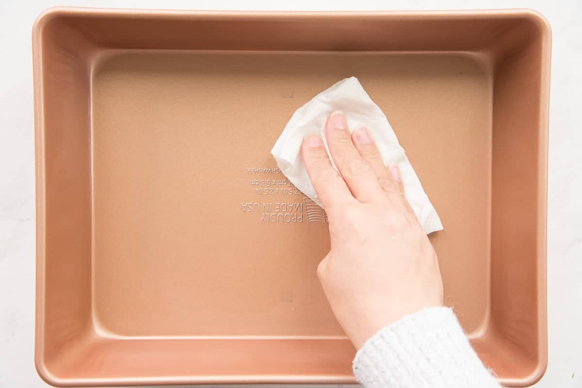 A hand lightly greases the bottom of a baking pan.
