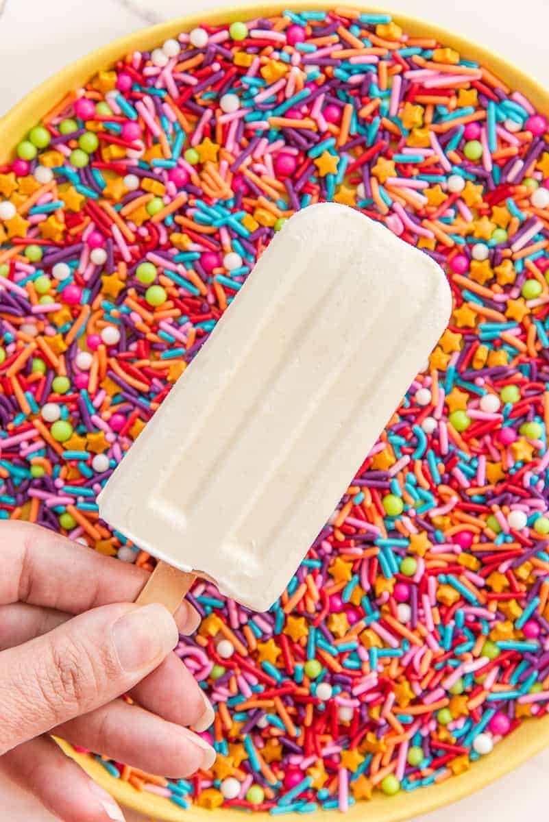A hand holds a tres leches paletas over a yellow plate of multicolored sprinkles.
