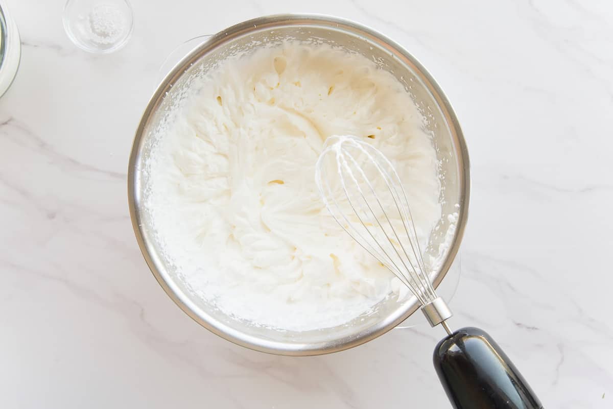 The whip of an immersion blender is lifted from the bowl of whipped cream.