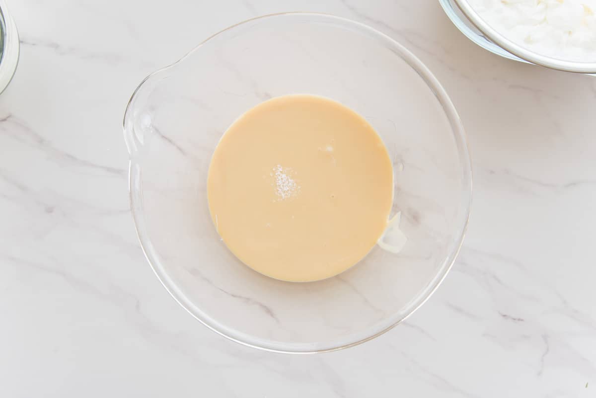 Salt and almond extract are added to the sweetened condensed milk in a glass bowl.