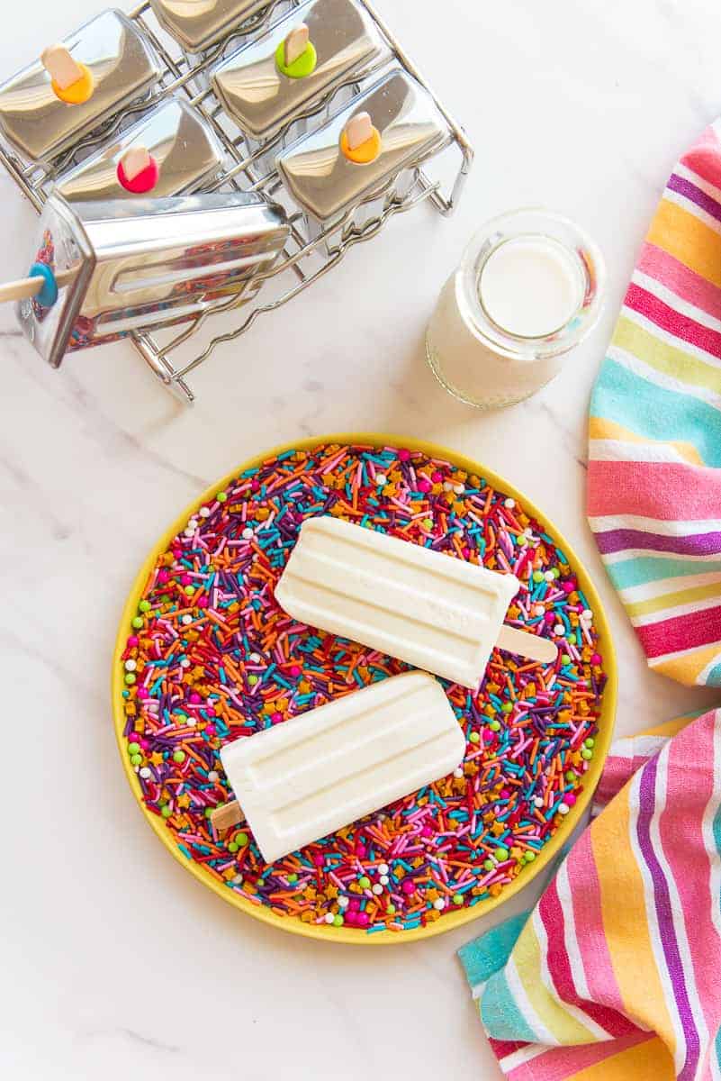 Two tres leches paletas on a yellow plate of rainbow sprinkles next to a bottle of milk and popsicle molds.
