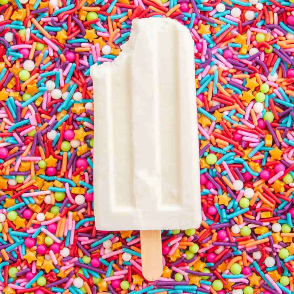 A tres leches paletas with a bite removed on a bed of multicolored sprinkles.