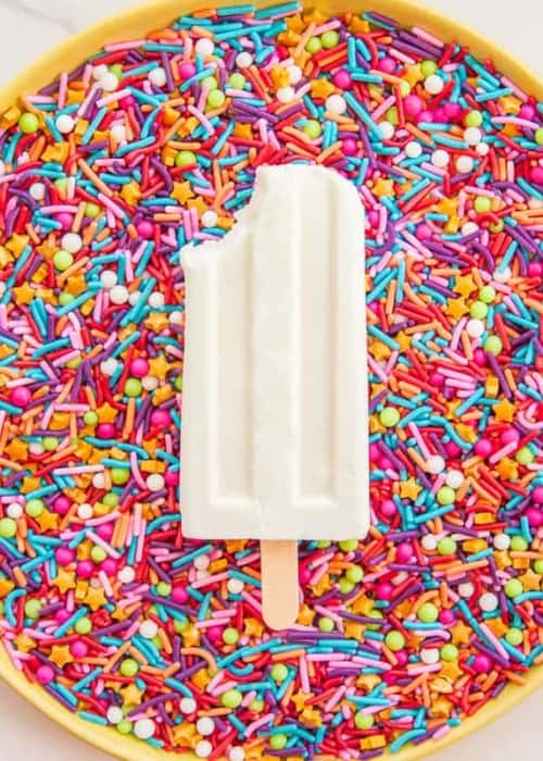 A tres leches paletas on a bed of rainbow sprinkles has a bite taken from it.