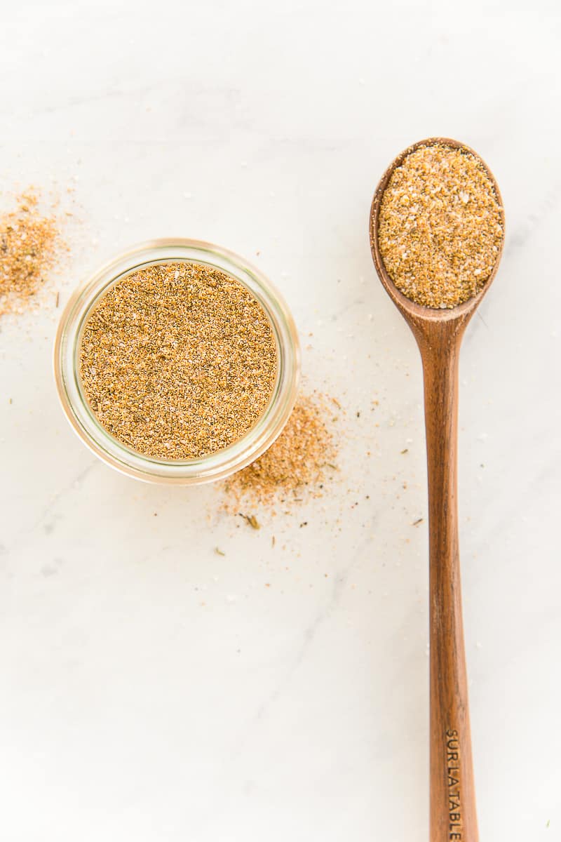 A glass jar of Chicken Seasoning Blend next to a dark wooden spoon of the spice blend