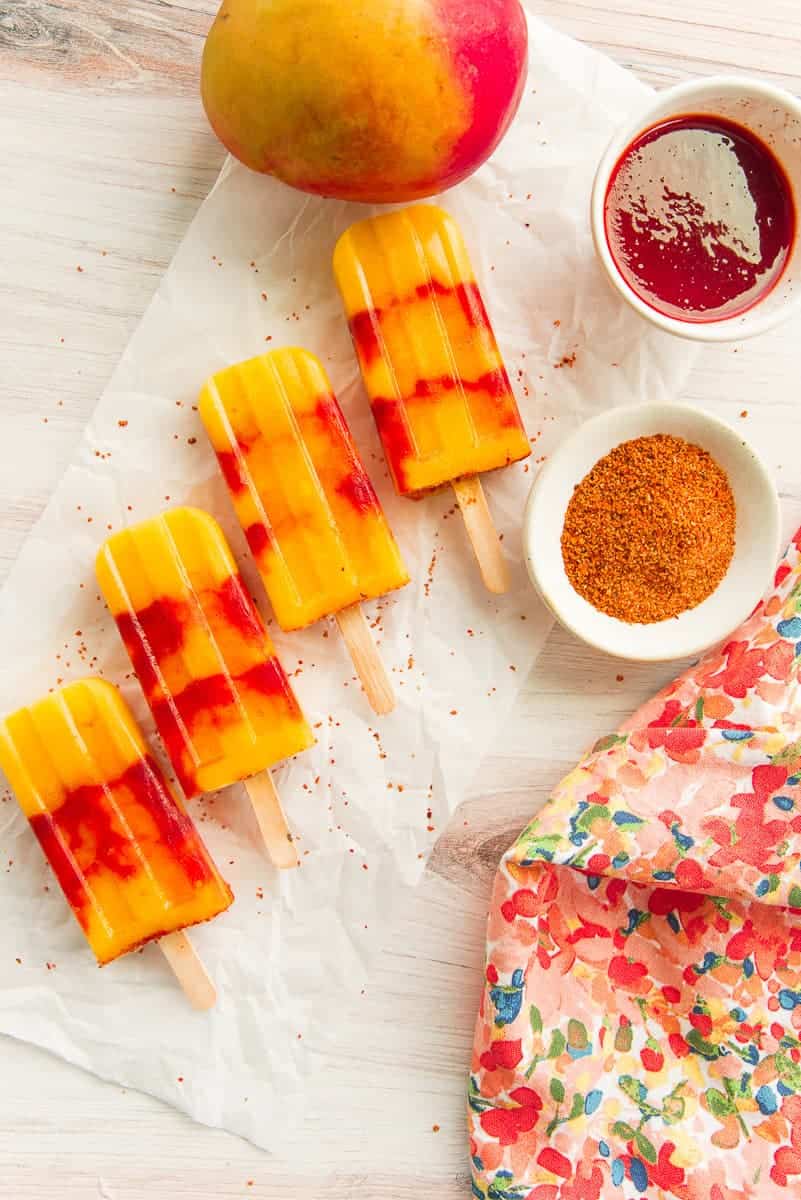 Mango Rosewater Paletas with Chamoy next to a ripe mango, a bowl of chamoy, and a bowl of tajín.