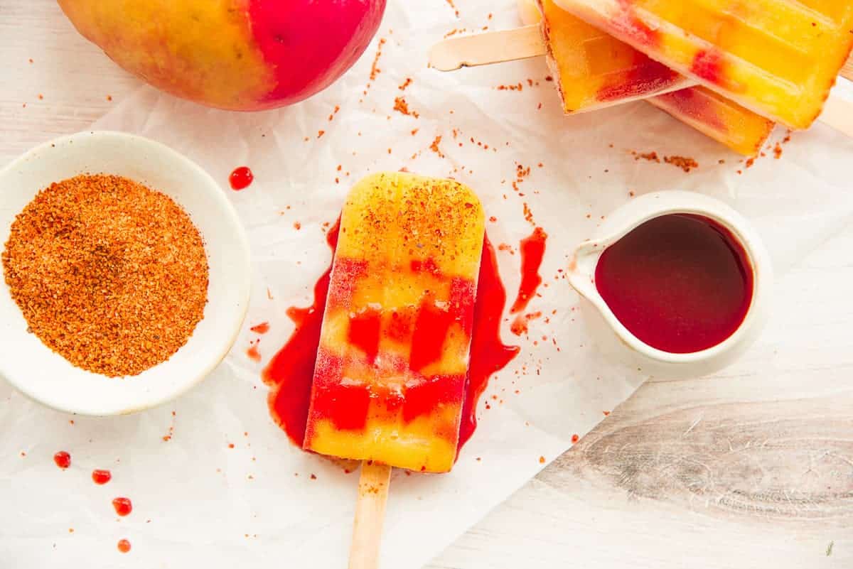 A single Mango Rosewater Paletas with Chamoy is drizzled with chamoy next to a bowl of tajin.