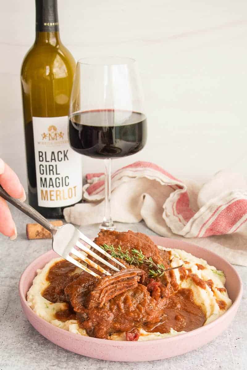 Merlot Braised Short Ribs in a pink bowl in front of a wine glass filled with merlot.