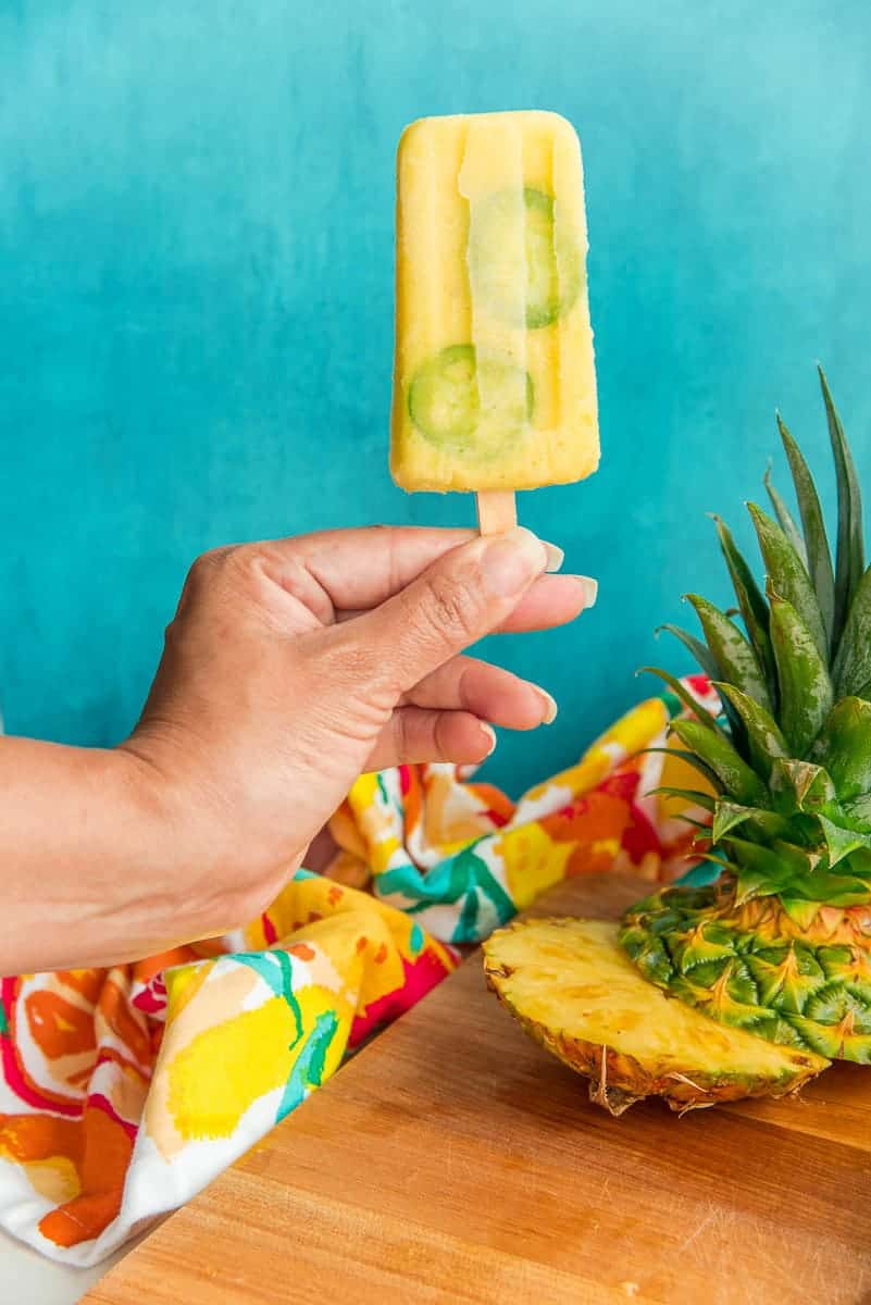 A hand holds a Spicy Pineapple Paletas in front of a blue background.