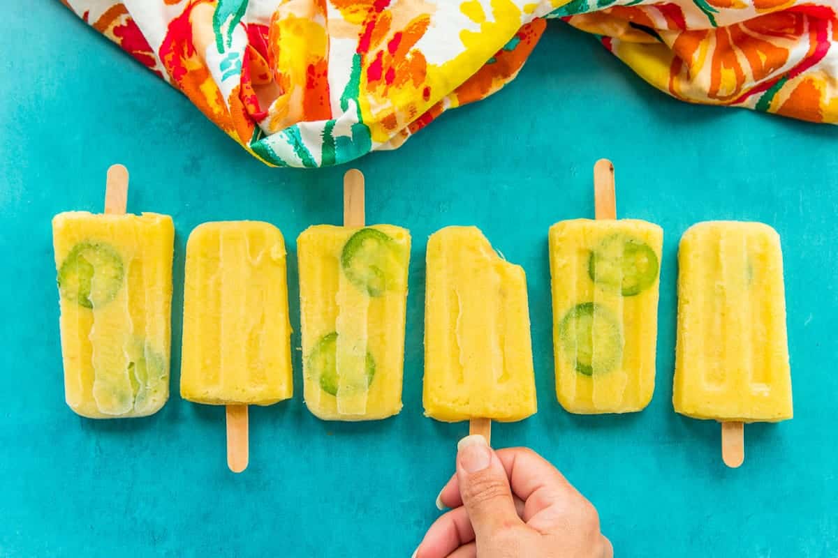 A hand reaches for a paleta from a line of Spicy Pineapple Paletas on a teal surface.