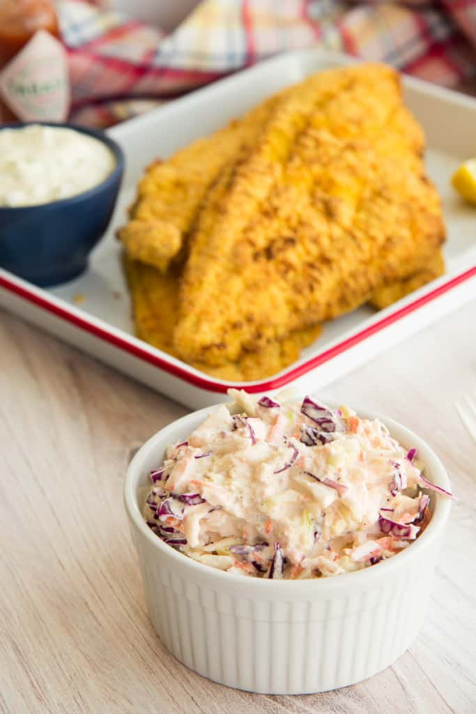A white dish filled with Homemade Coleslaw in front of a tray with fried fish on it.
