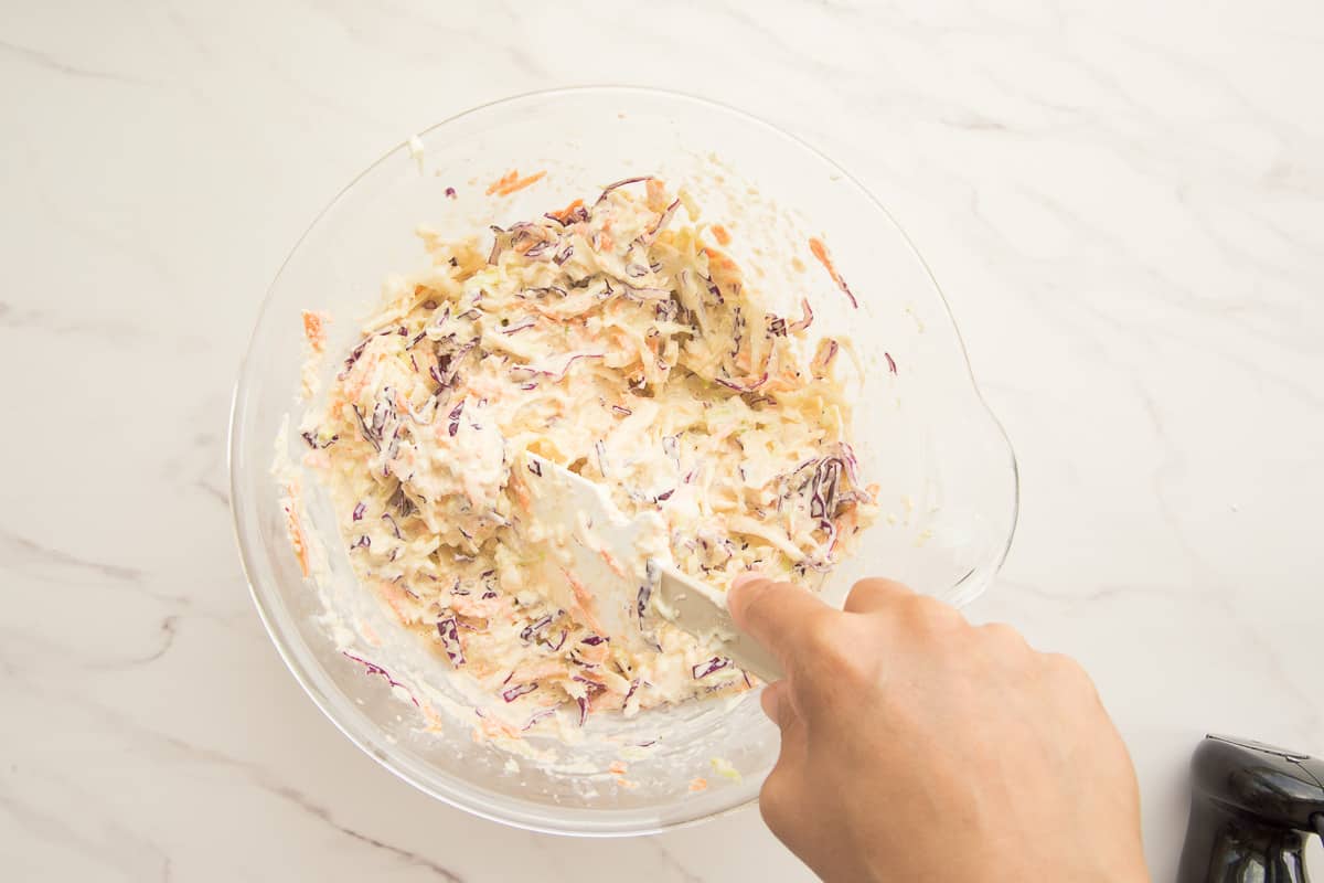 A hand using a rubber spatula to stir the dressing into the shredded veggies in a clear bowl.