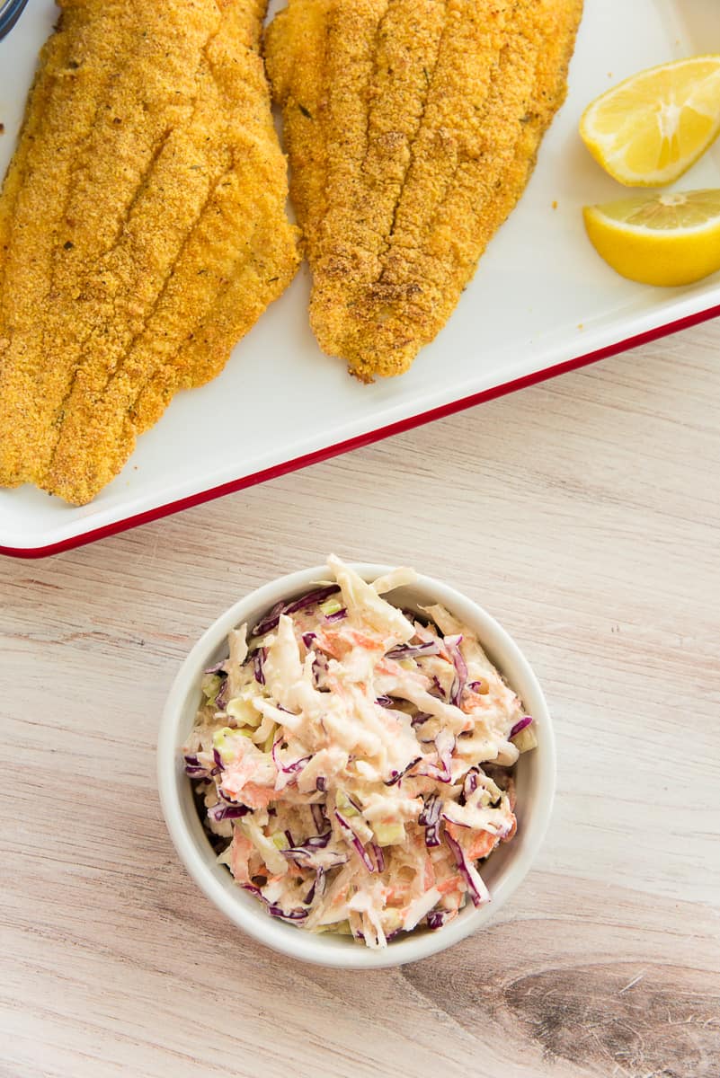 A white ramekin of Homemade Coleslaw next to a tray with fried fish and lemon wedges on it.