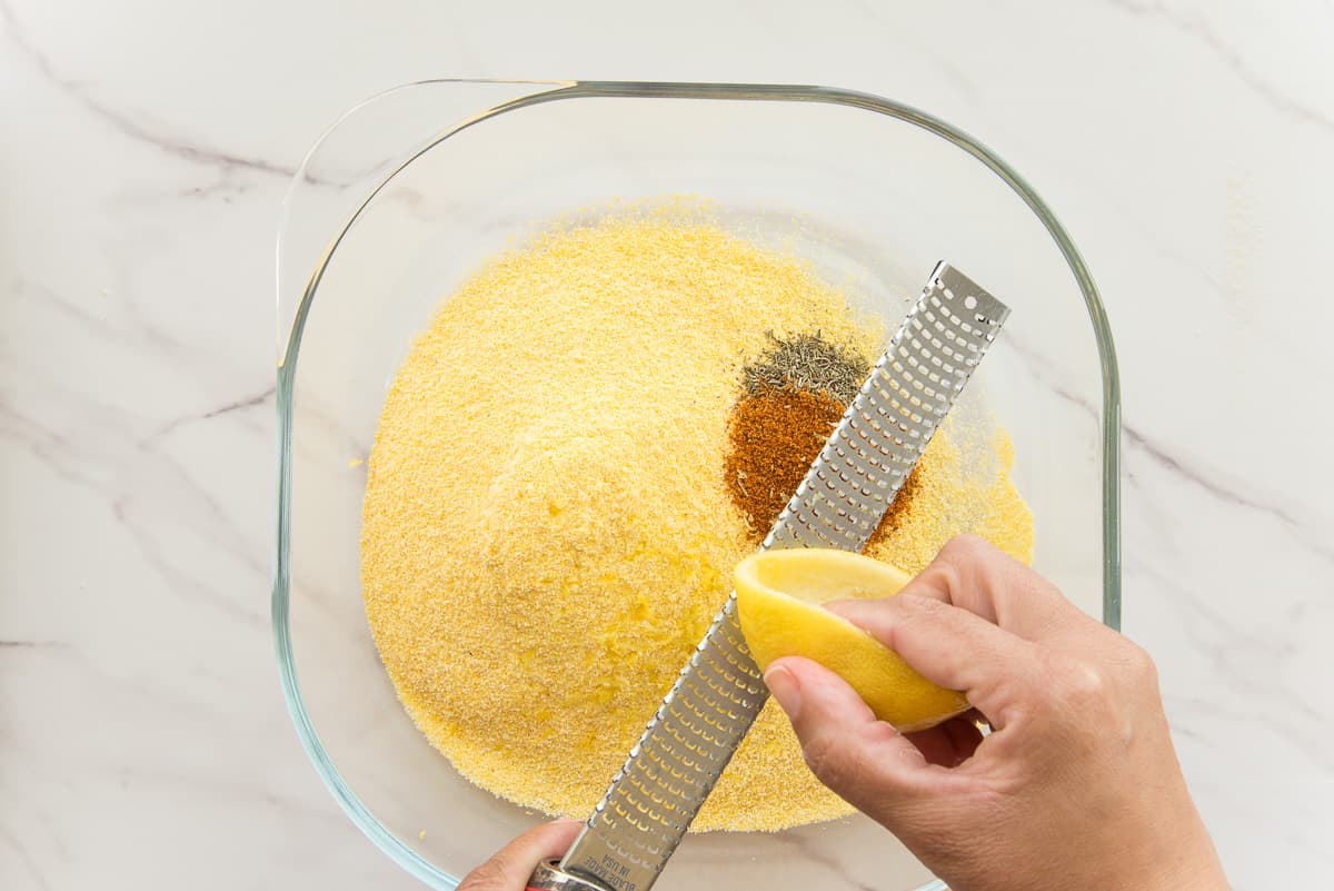 A hand uses a microplane to zest the lemon peel into cornmeal with piles of spices on it for the Air Fried Southern Catfish