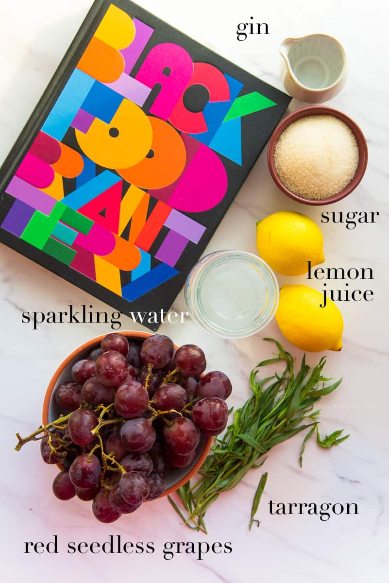 Ingredients to make the Grape Tarragon Gin Spritzer on a white surface next to Bryant Terry's Black Food Cookbook.