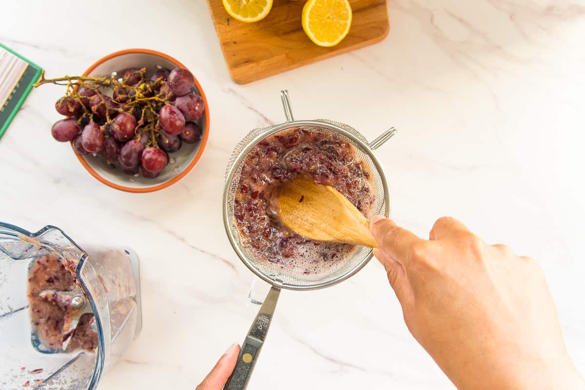 A hand uses a wooden spoon to press the juice from blended grapes.
