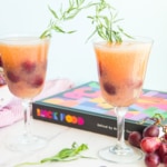 Two Grape Tarragon Gin Spritzers in goblets are garnished with tarragon sprigs in front of Black Food Cookbook.